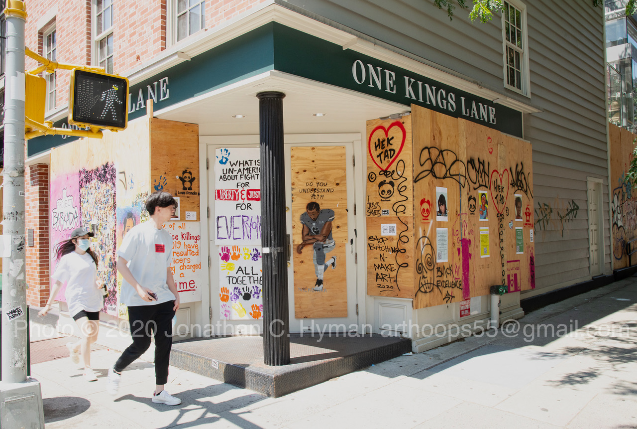 Plywood-boarded storefront covered in hand-painted Black Lives Matter street art with pedestrians walking past