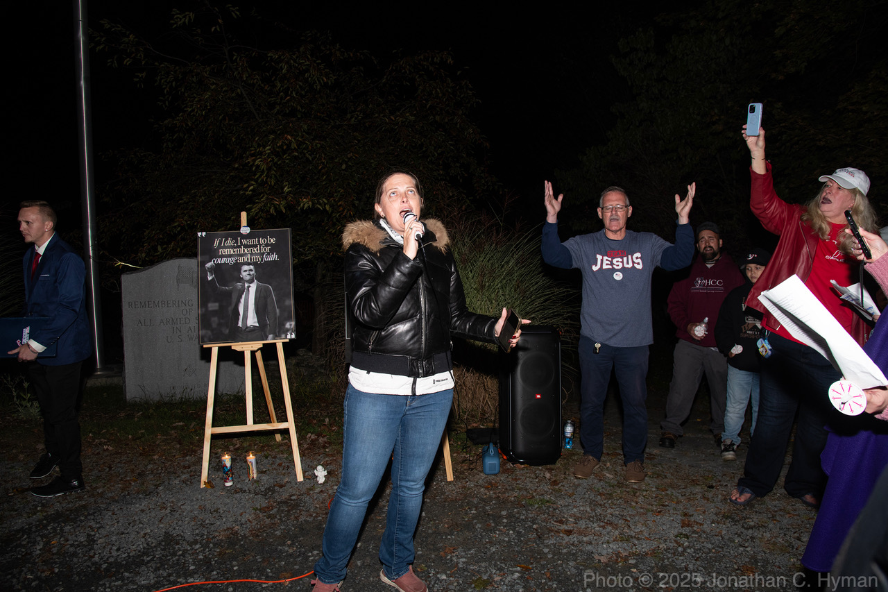 A woman singing into a microphone at a nighttime candlelight vigil beside a framed portrait of Charlie Kirk