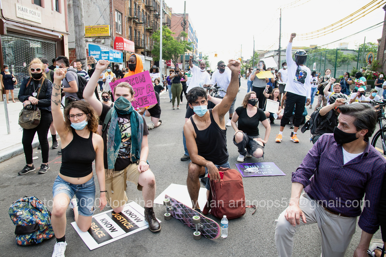 Masked protesters kneeling with raised fists in a Brooklyn street beside hand-lettered protest signs