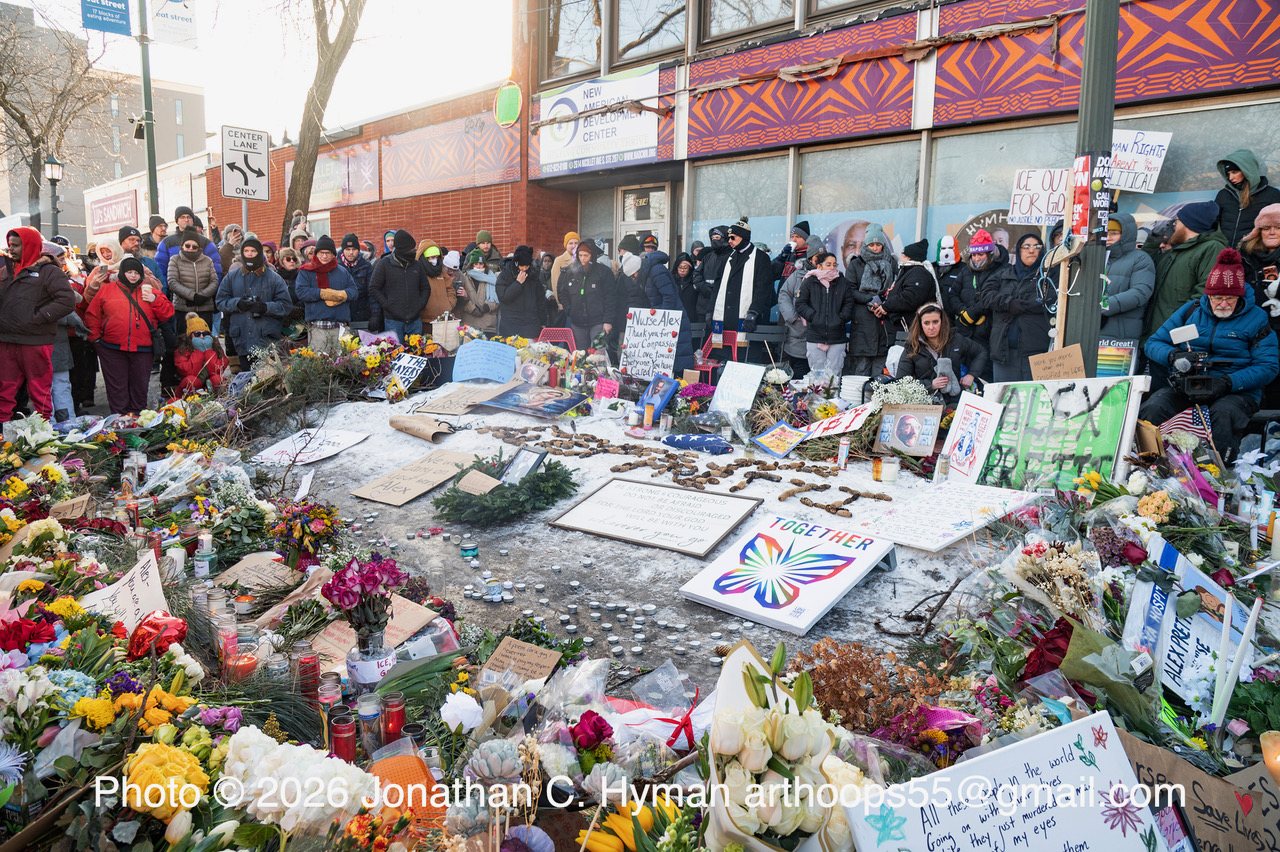 A large sidewalk memorial of flowers, candles, and hand-written signs with mourners gathered around it in the snow