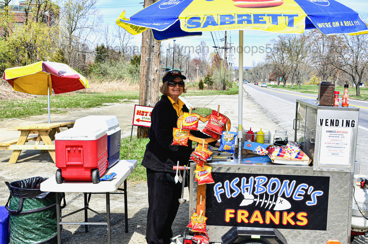 Vendor standing beside the Fishbone's Franks roadside hot dog cart under a Sabrett umbrella near Pawling, New York