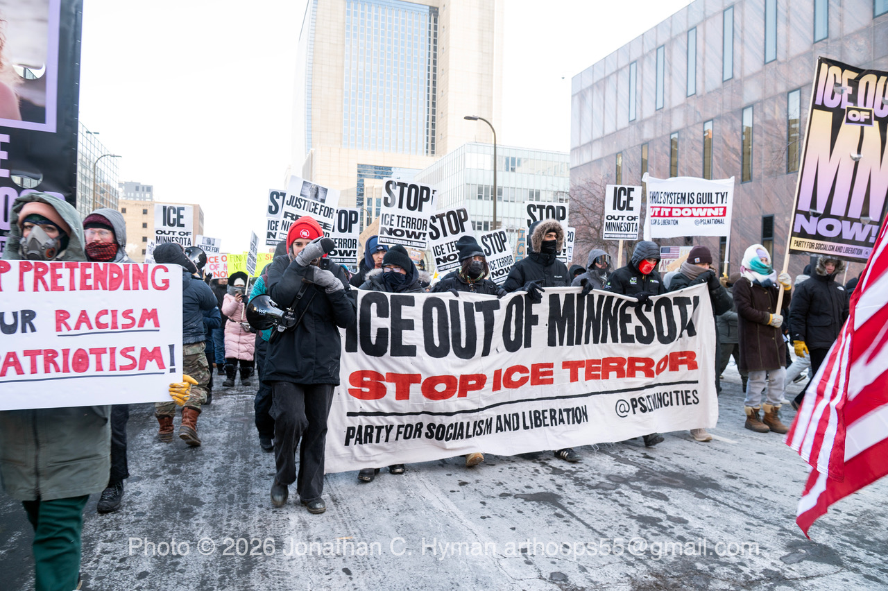 Marchers in winter clothing carrying an Ice Out of Minnesota banner and protest signs through a snowy downtown street