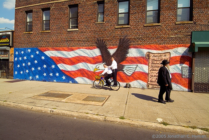 Two boys riding a bicycle past a hand-painted eagle and American flag mural on a Brooklyn brick wall