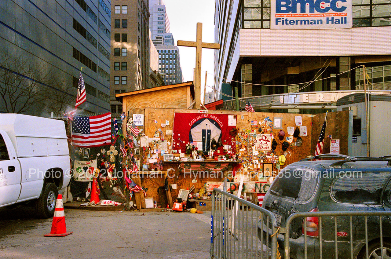 Wooden cross above a plywood roadside memorial covered with flags, patches, and tributes near Ground Zero in Lower Manhattan