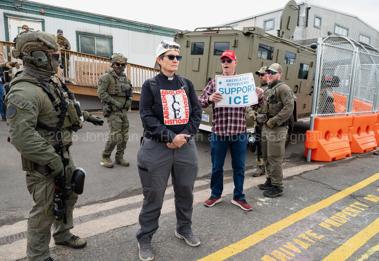 A woman clergy member standing between armed ICE agents and a counter-protester holding a 'Support ICE' sign outside Delaney Hall in Newark, New Jersey