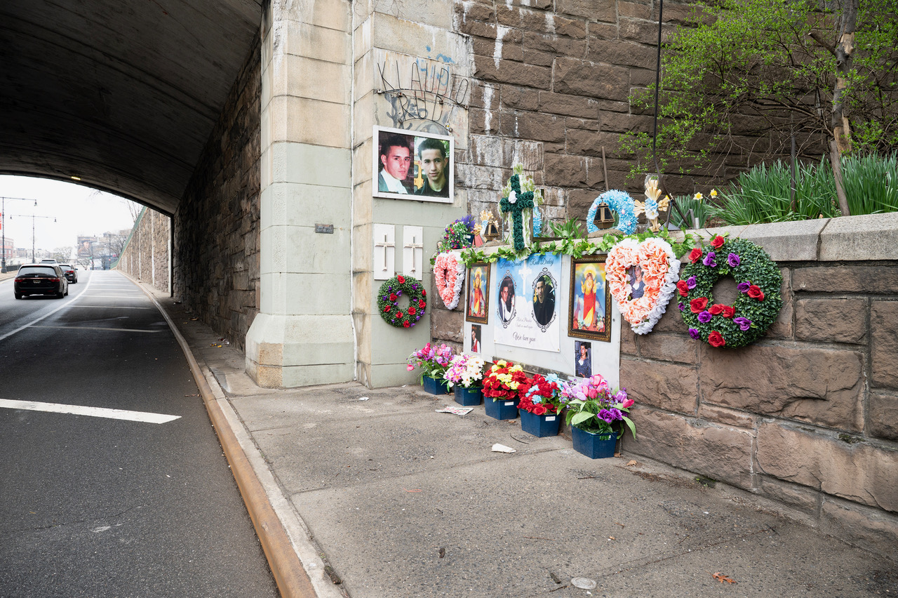 A roadside memorial under a stone bridge in the Bronx with photographs, crosses, wreaths, and flowers