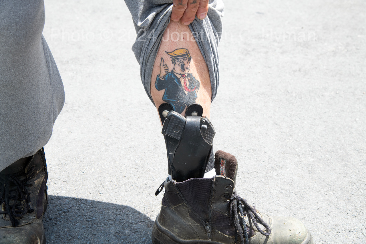 A man lifting his pant leg to reveal a cartoon Donald Trump tattoo above a holstered handgun tucked into his boot